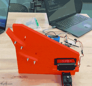 A bright orange robotic component with wires, placed on a wooden desk alongside laptops.