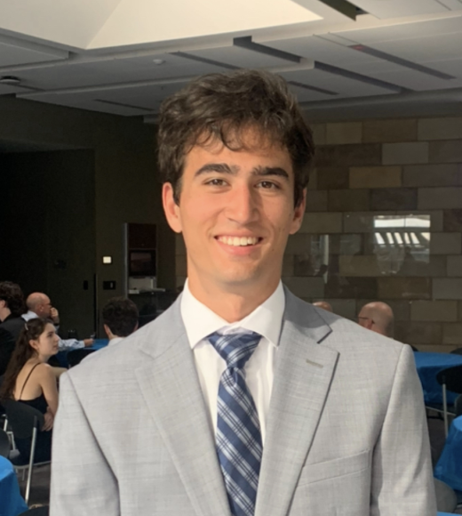 A young man in a light gray suit smiles, standing in a room with round tables.