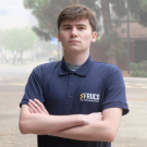 A young man stands confidently outdoors, arms crossed, wearing a navy polo shirt.